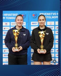 Two female athletes stand side by side, each holding a tall gold trophy, smiling in front of a blue sponsor backdrop.