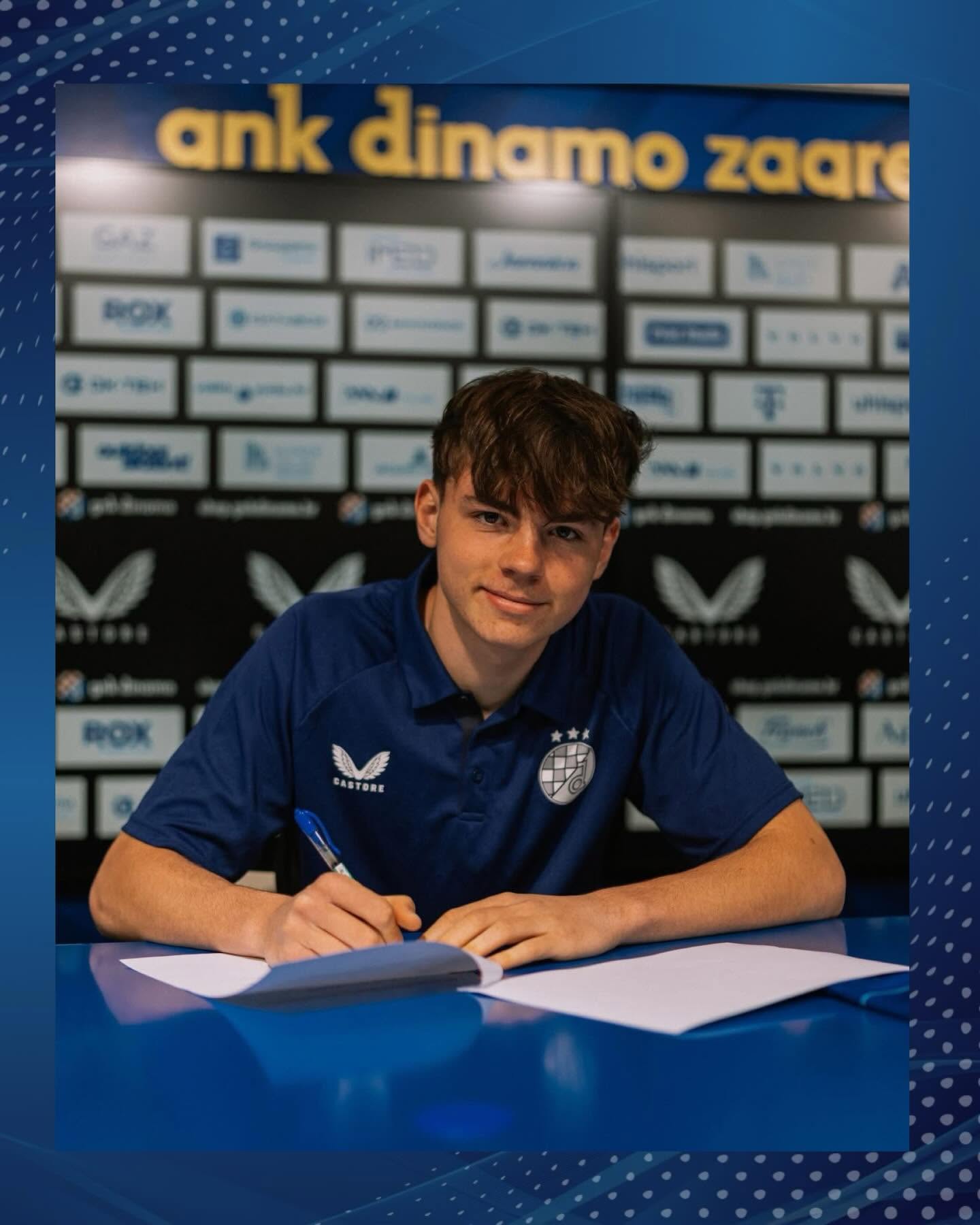 Young man in a blue polo signs a document at a table, backdrop filled with sponsor logos behind him