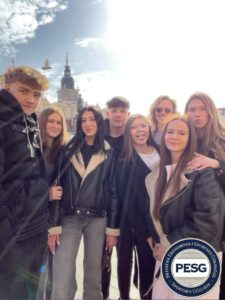 Group of eight young people posing for a selfie on a sunny day, wearing jackets, with a historic building in the background and a PESG logo in the bottom right corner.