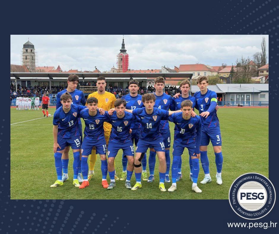 Youth soccer team in blue uniforms poses for a group photo on a grassy field, with the goalkeeper in yellow at the back center and a stadium in the background.