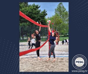 Two women playing beach volleyball outdoors, reaching for a ball over a red net with onlookers in the background; PESG logo bottom right.
