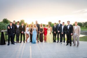 Group of teens in formal dresses and suits posing outdoors at sunset on a tiled terrace, with a green lawn and pond in the background.