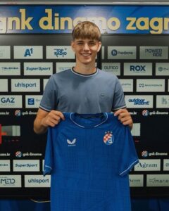 Young man wearing a light blue shirt holds up a blue soccer jersey with a checkerboard crest, standing in front of a sponsor-filled backdrop.