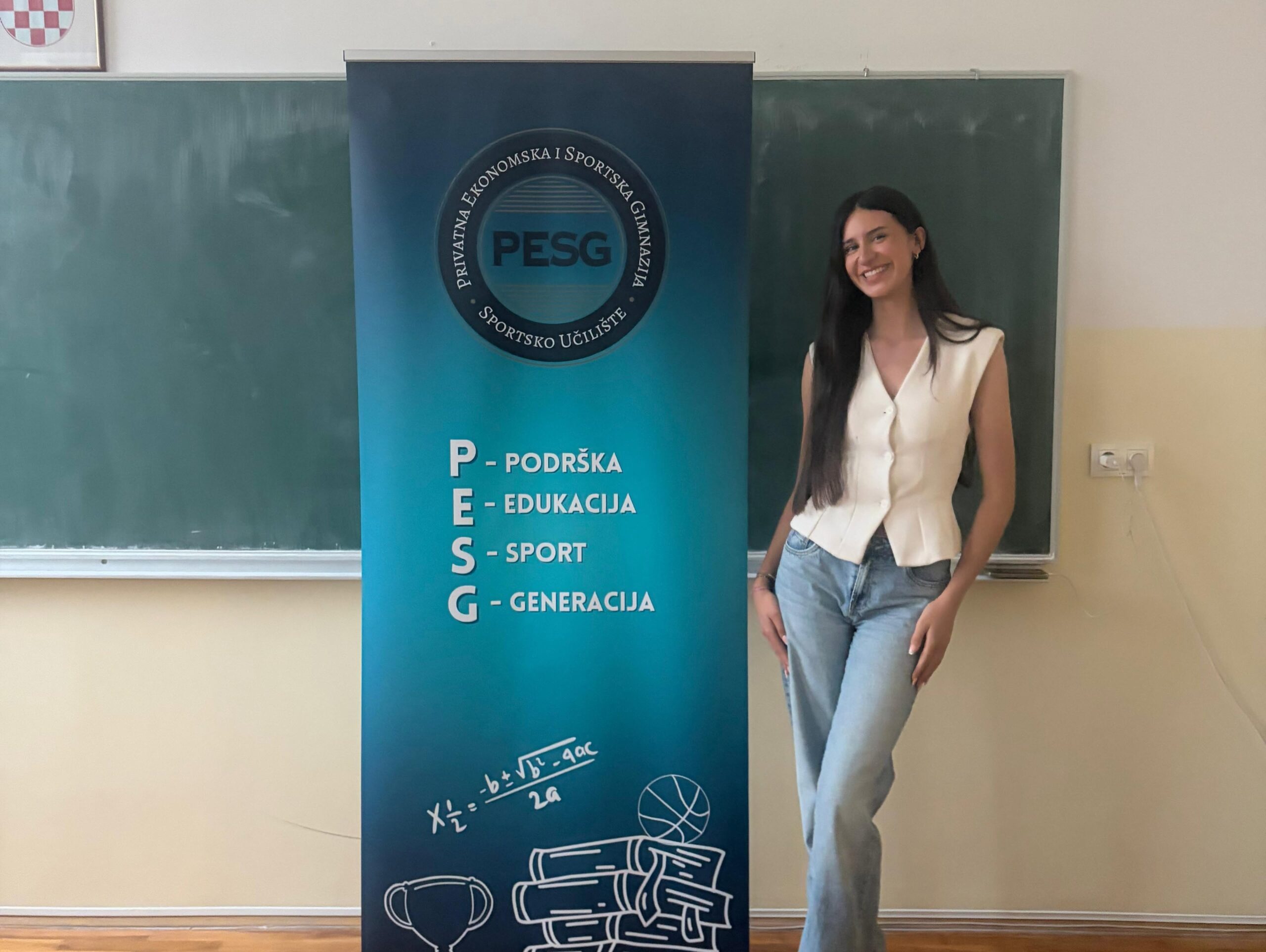 Smiling woman in a white top and jeans standing beside a blue PESG banner in a classroom crest logo visible on banner.