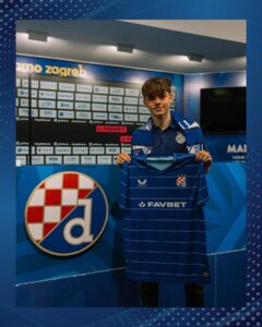 Young soccer player in a blue Dinamo Zagreb kit holding up a matching jersey in a team press area with sponsor boards behind him.