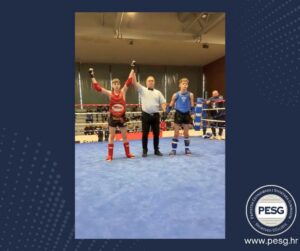 Two young boxers in red and blue gear stand with arms raised by a referee inside a boxing ring, signaling the match outcome.