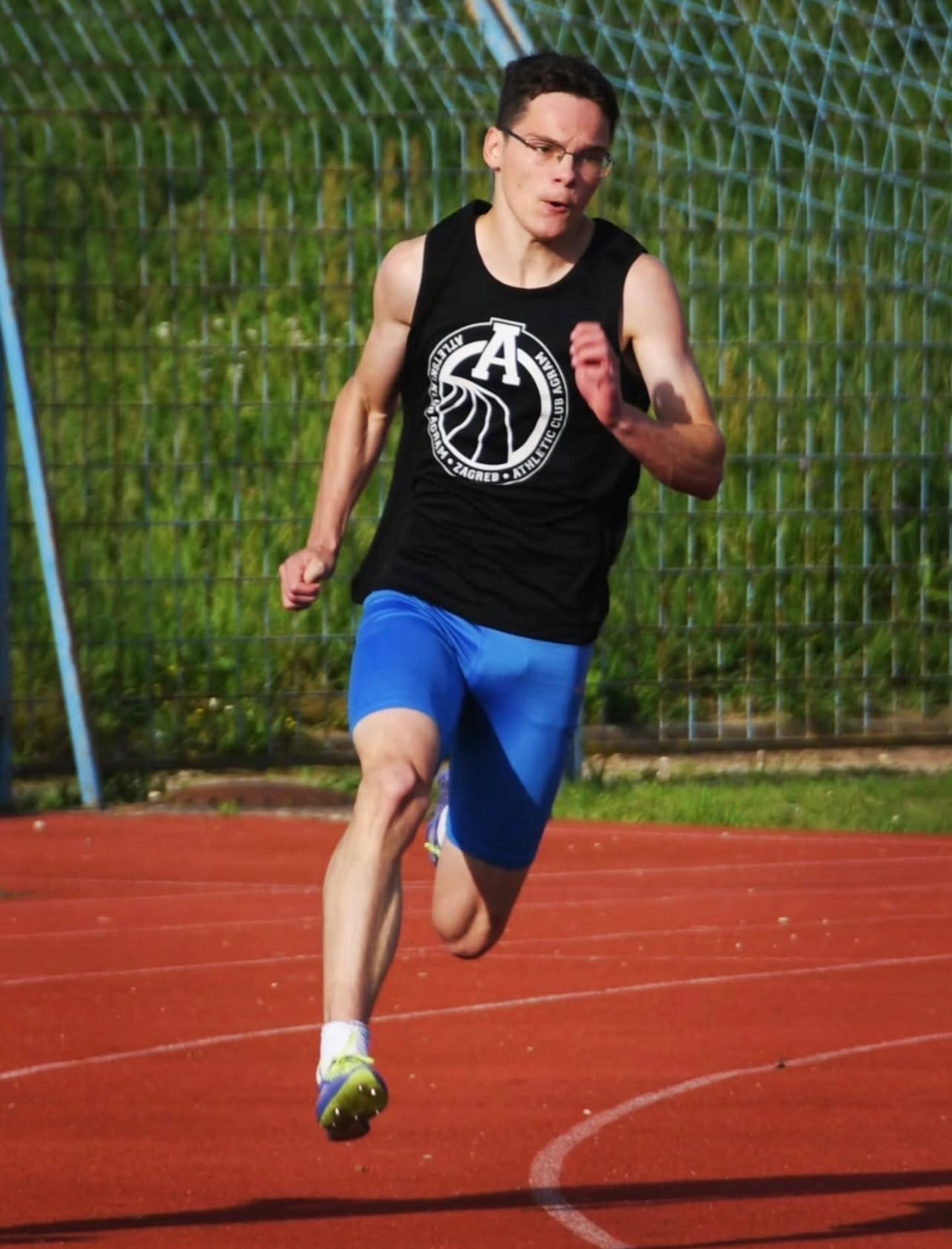 Male runner in a black sleeveless shirt and blue shorts sprinting on a red track; a chain-link fence and greenery are in the background.