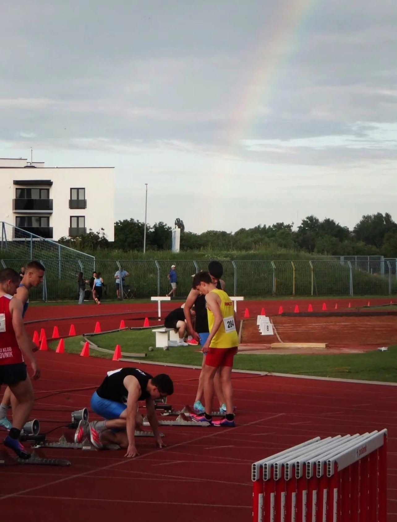 Track and field athletes prepare at a red running track with hurdles and a rainbow in the sky behind them.