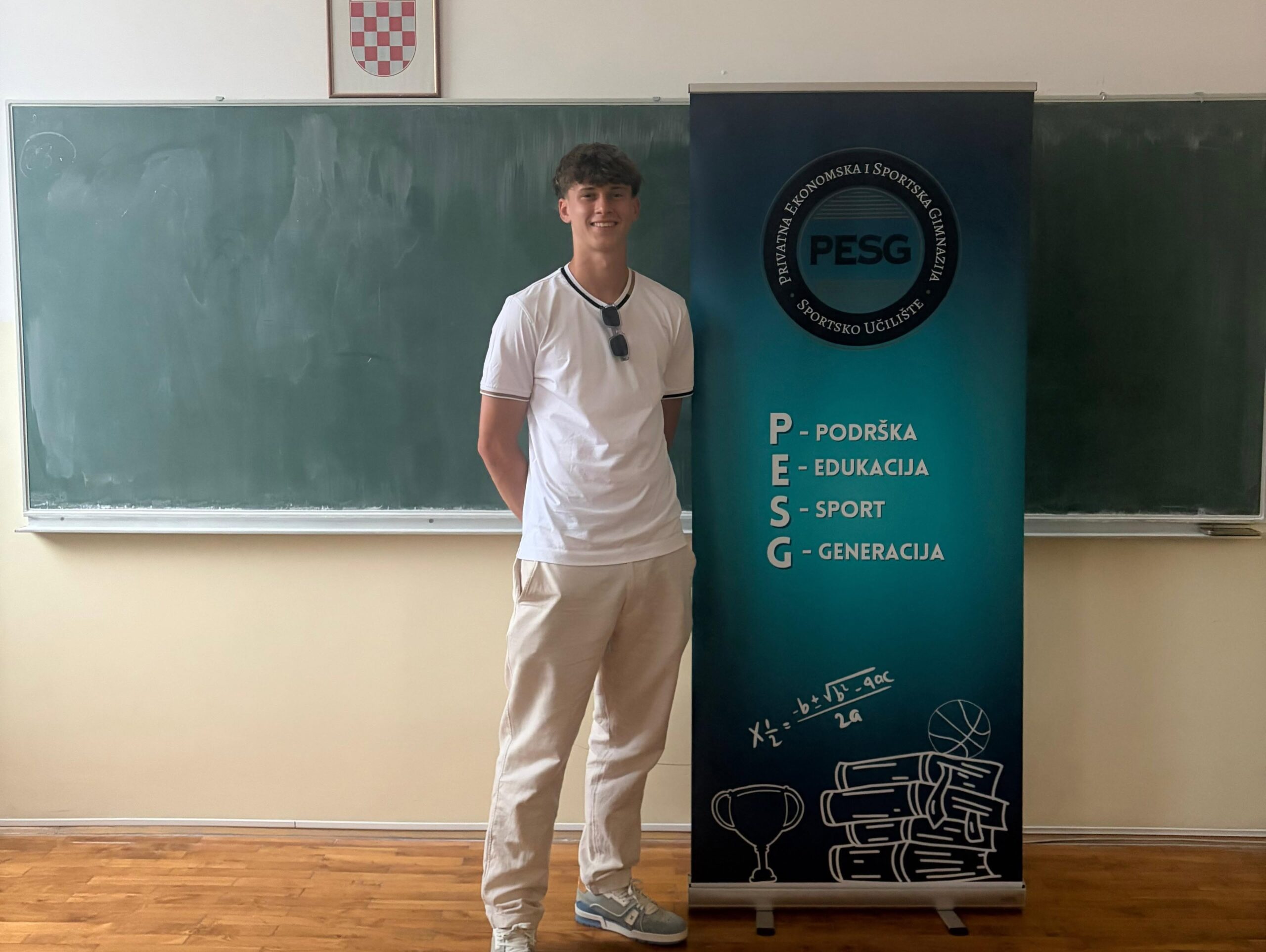Young man in a white shirt and light pants stands beside a blue PESG banner in a classroom, smiling.
