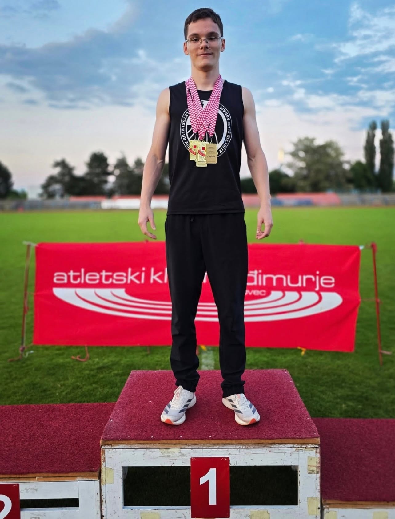 Young man on a first-place podium wearing a black sleeveless shirt and multiple medals, outdoors on a track field.