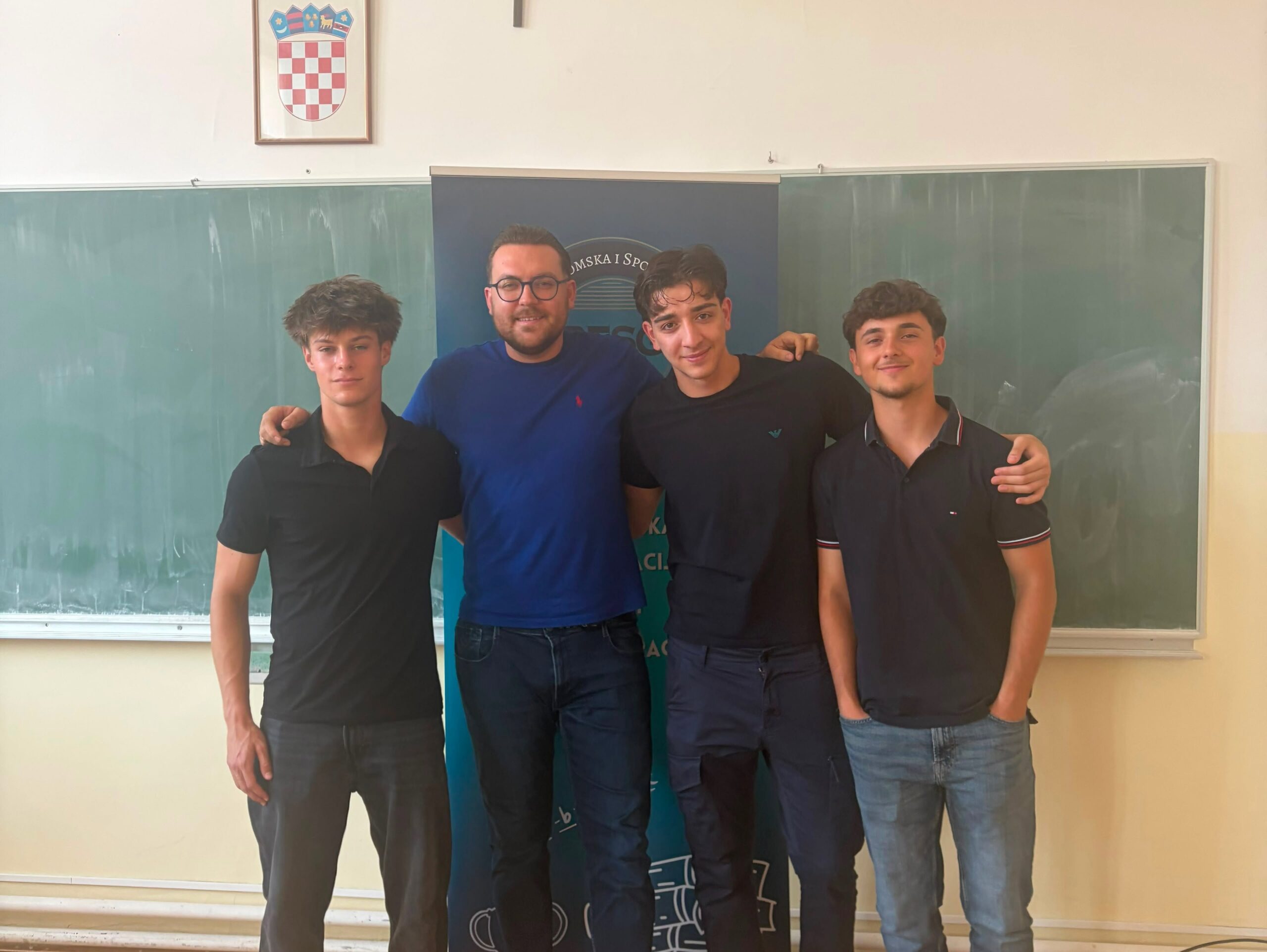 Four young men stand together in a classroom, arms around each other, in front of a banner and chalkboards.