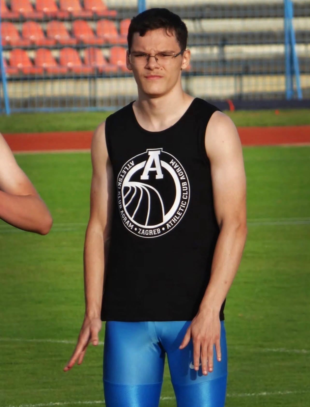 Young man in a black sleeveless athletic shirt and blue shorts standing on a track field, with empty stadium seats in the background.