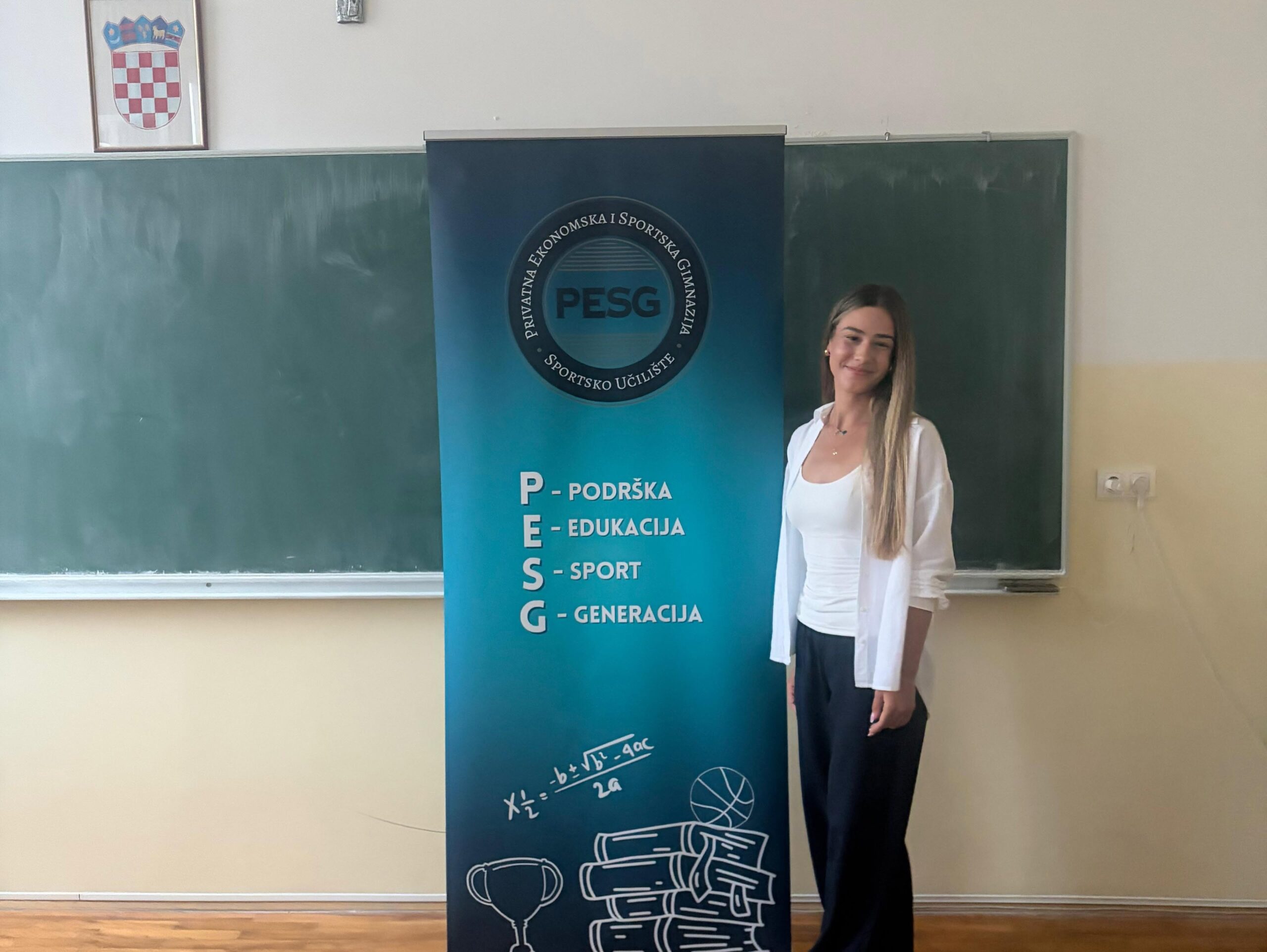Banner for PESG in a classroom, with a smiling woman standing beside it and a chalkboard in the background.
