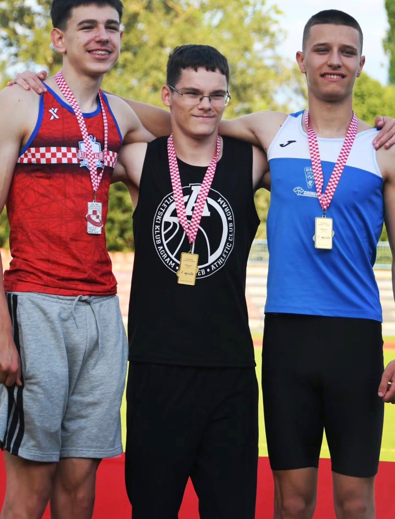 Three young male athletes pose with medals after a race, arms around each other on a track.