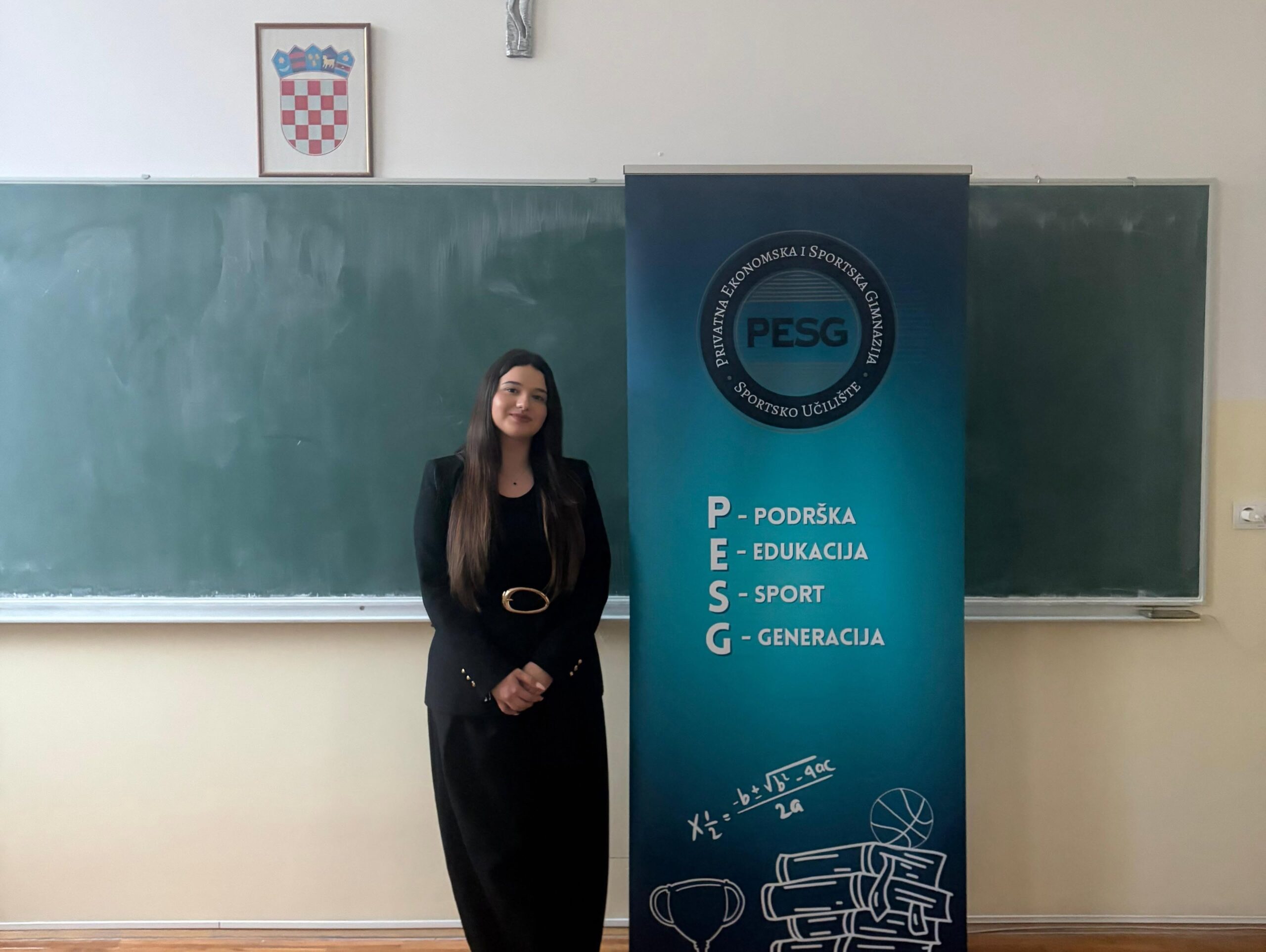 Young woman in a black suit standing beside a blue PESG banner in a classroom setting. (informative)