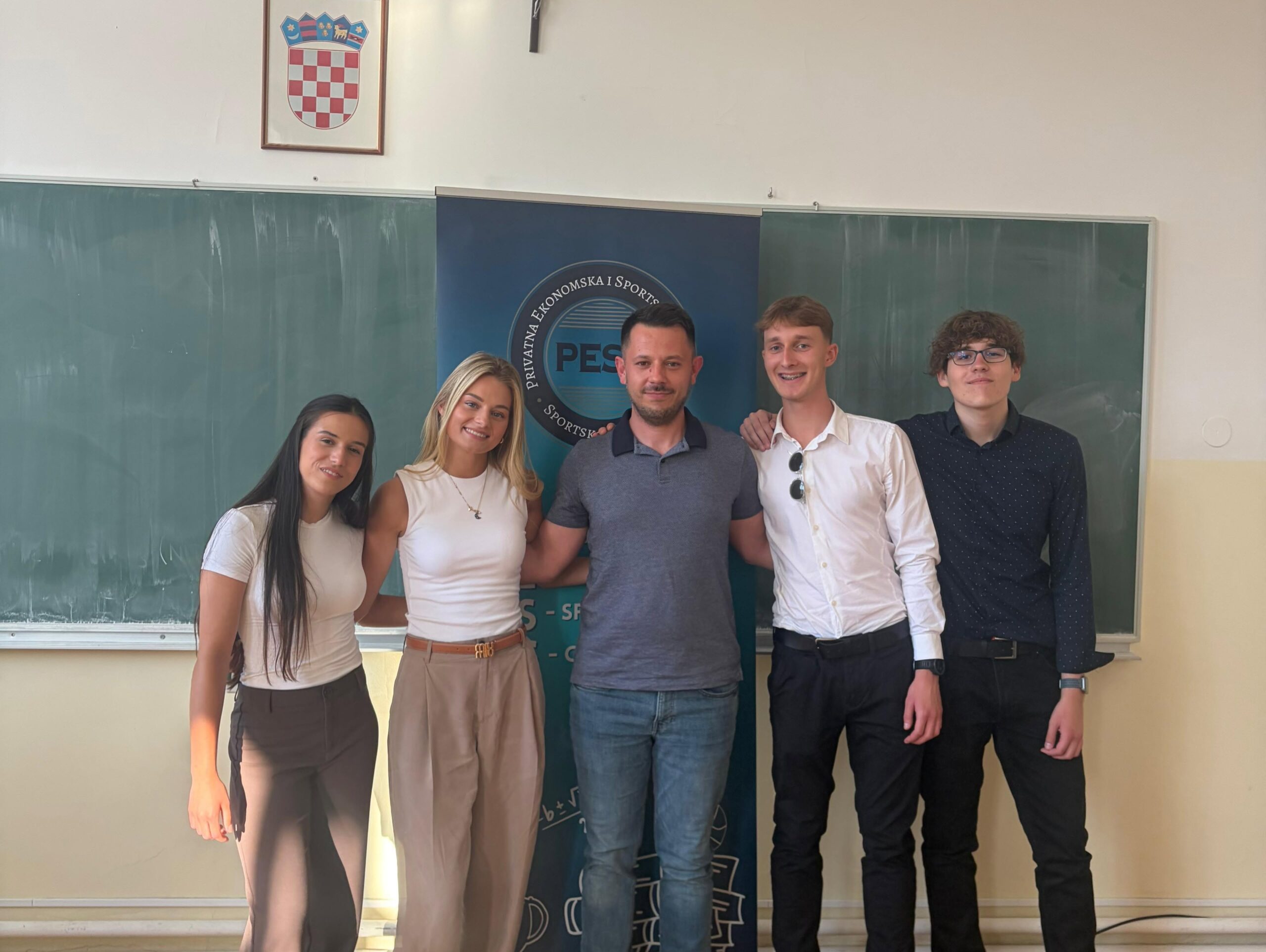 Five students posing in a classroom in front of a blue banner with a circular logo, chalkboard behind, Croatian coat of arms on the wall above.