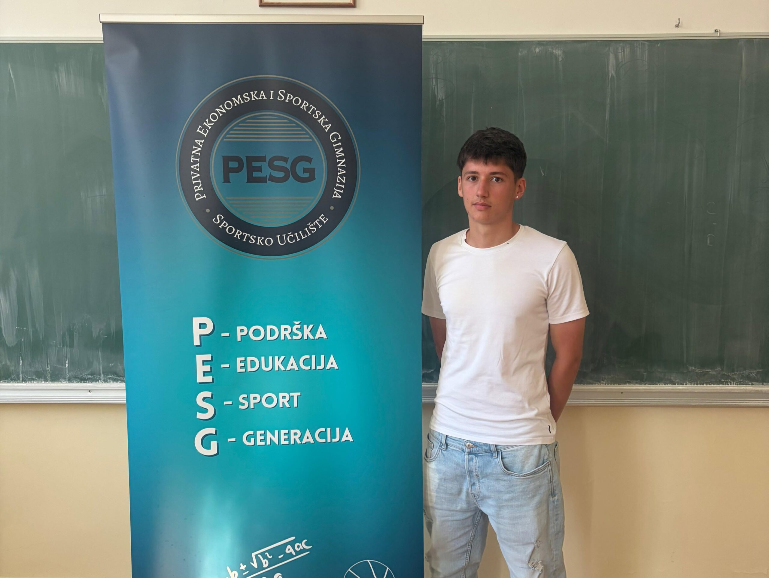 Young man in a white t-shirt stands beside a blue PESG banner displaying the logo and a vertical list of topics (Podrška, Edukacija, Sport, Generacija).