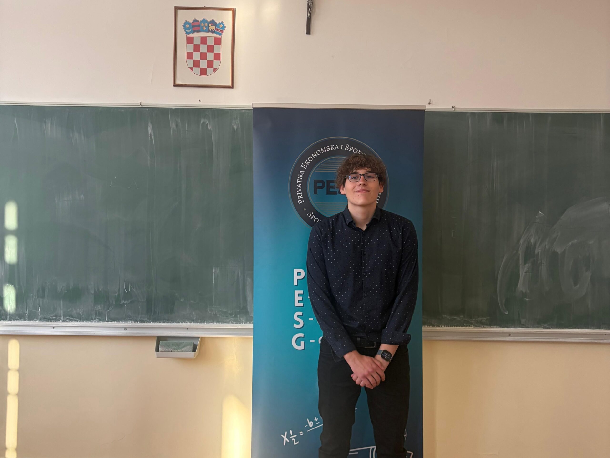 Young man with glasses standing in a classroom, in front of a blue banner with a circular logo and a chalkboard backdrop.