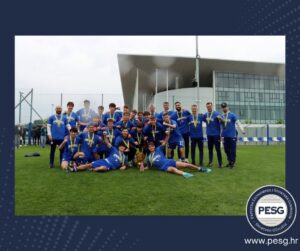 Group of young male soccer players in blue uniforms posing with medals and a trophy on a green field, with a modern building in the background; PESG logo bottom right.