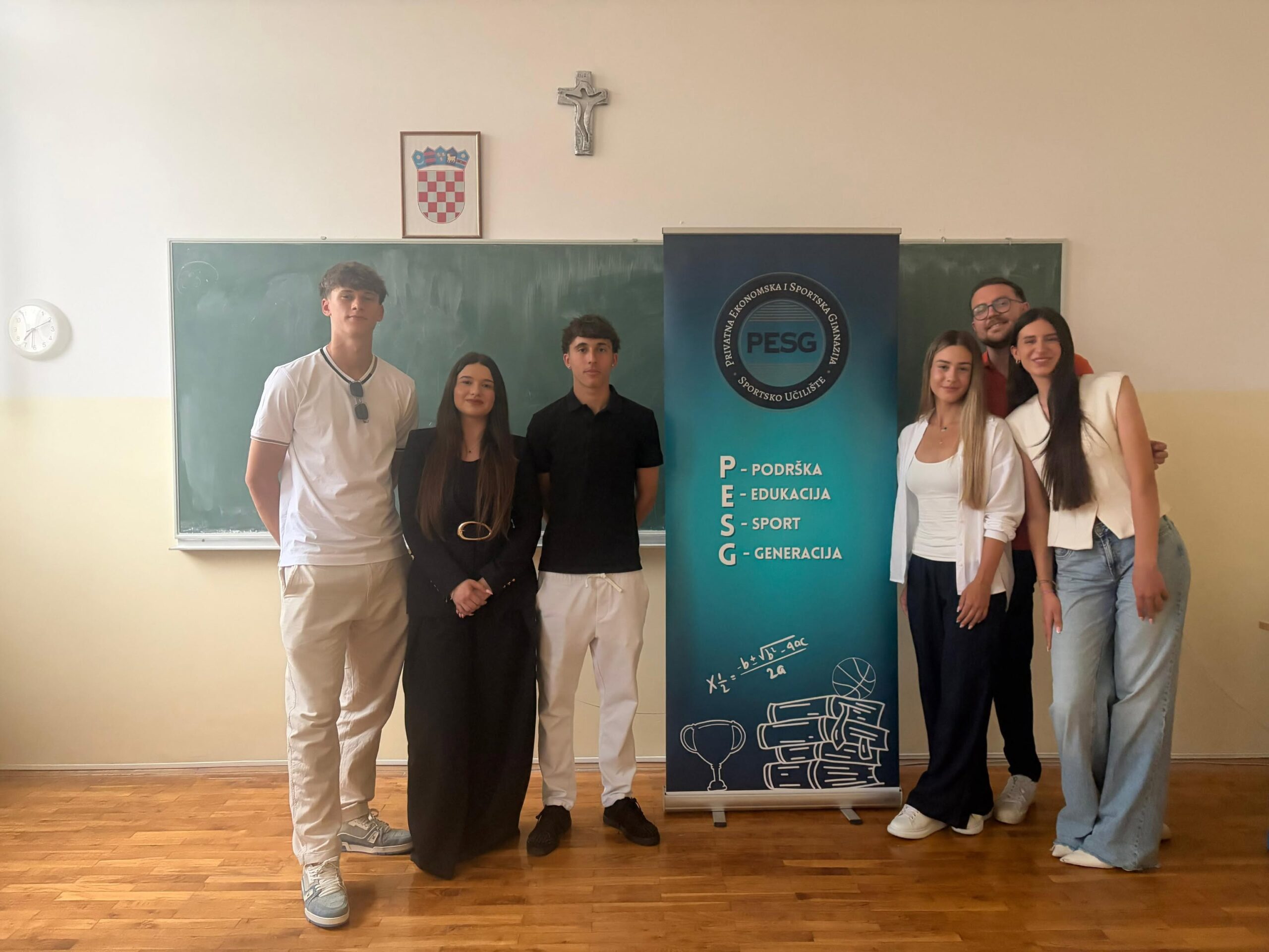 Group of six students posing in a classroom beside a PESG banner, with a chalkboard and a crucifix on the wall behind them.