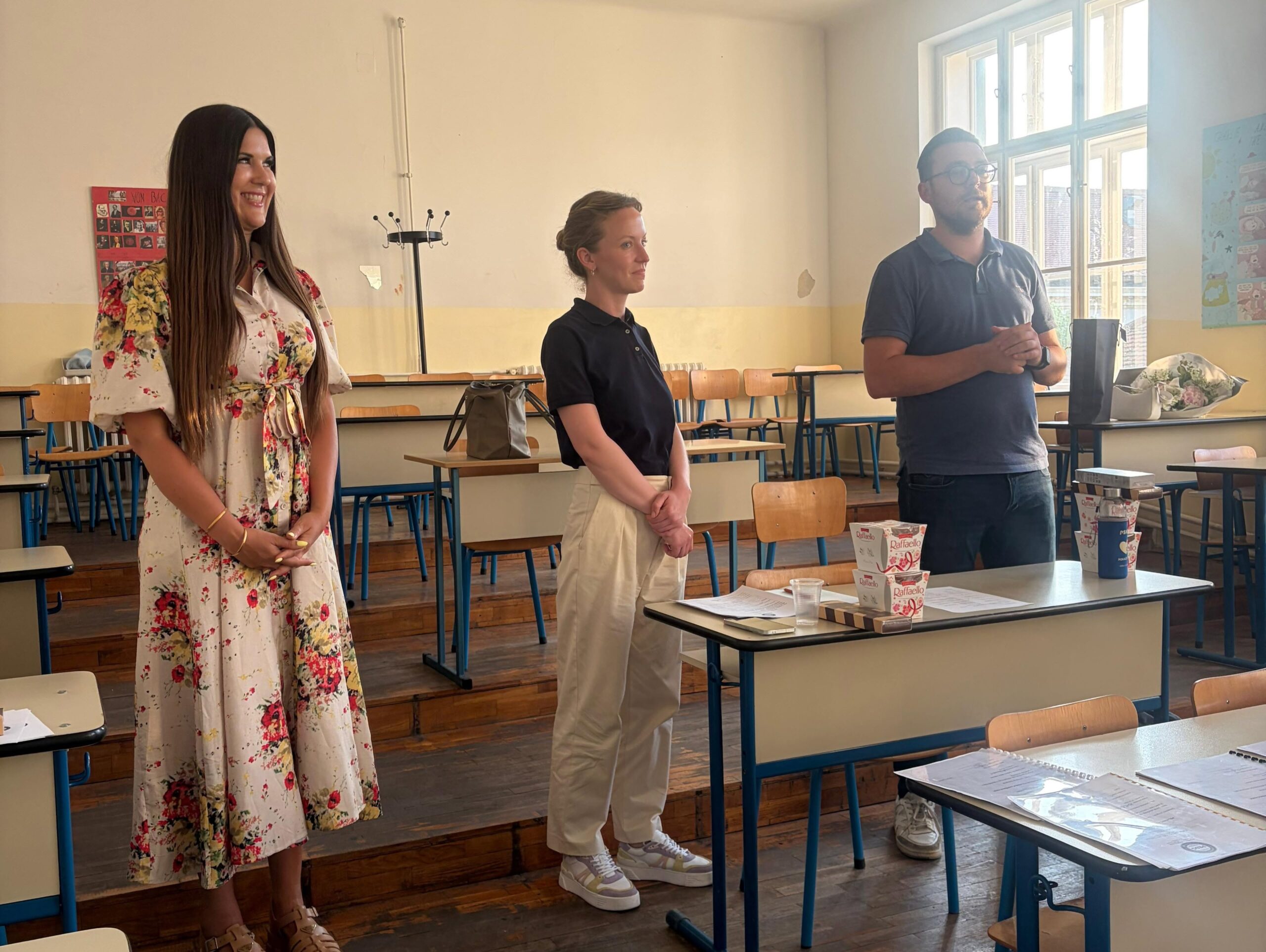 Three adults stand at the front of a classroom, smiling, with desks and chairs around them.