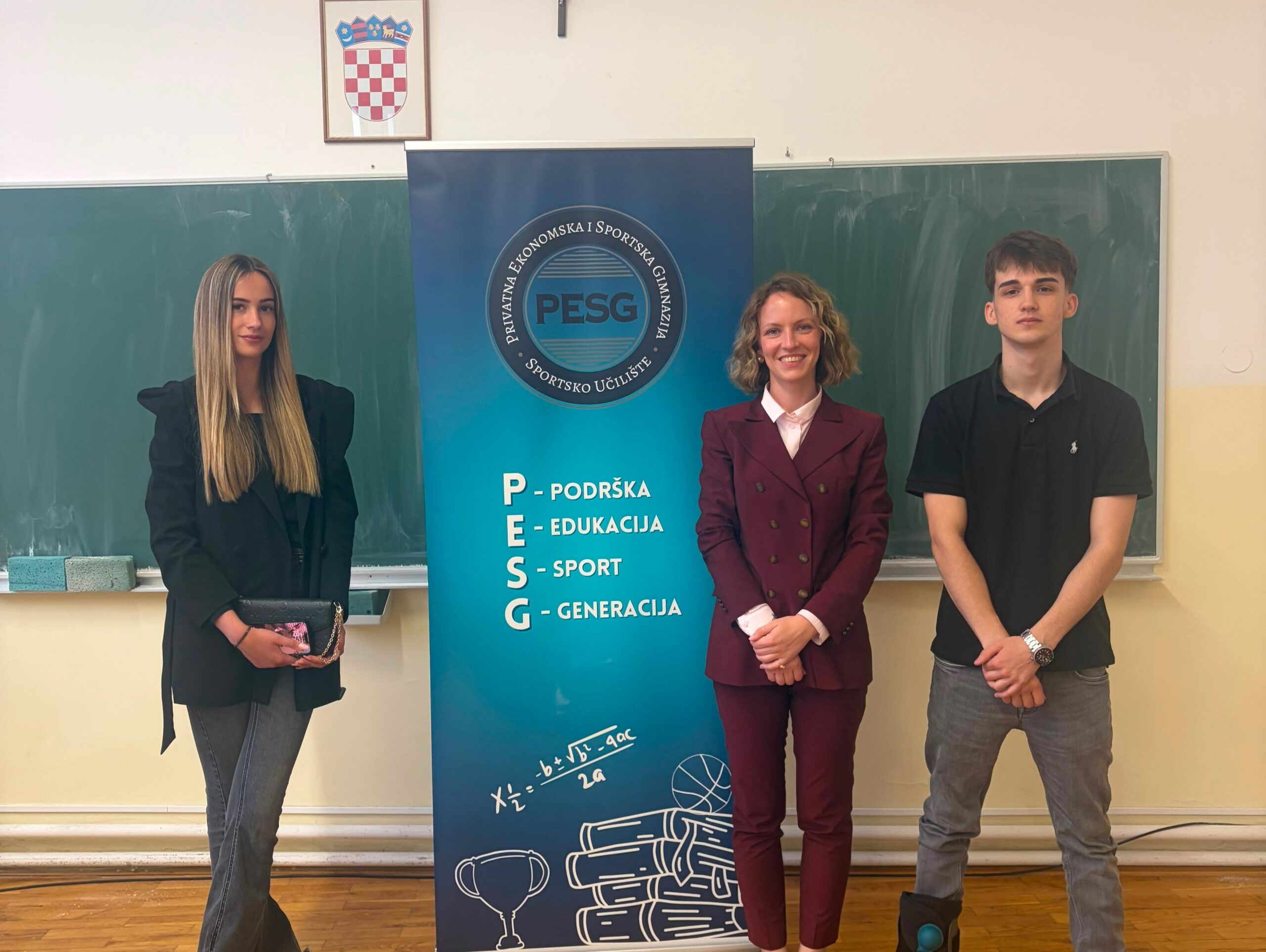 Three students posing beside a tall blue PESG banner in a classroom, with the PESG acronym and mission words visible.
