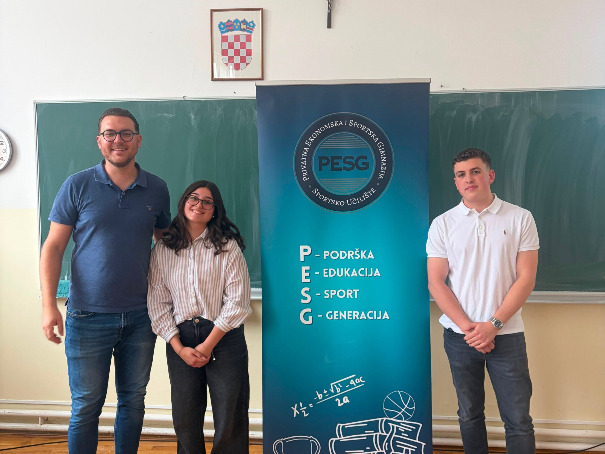 Three students pose beside a blue PESG banner in a classroom, with a crest on the banner and a chalkboard in the background.