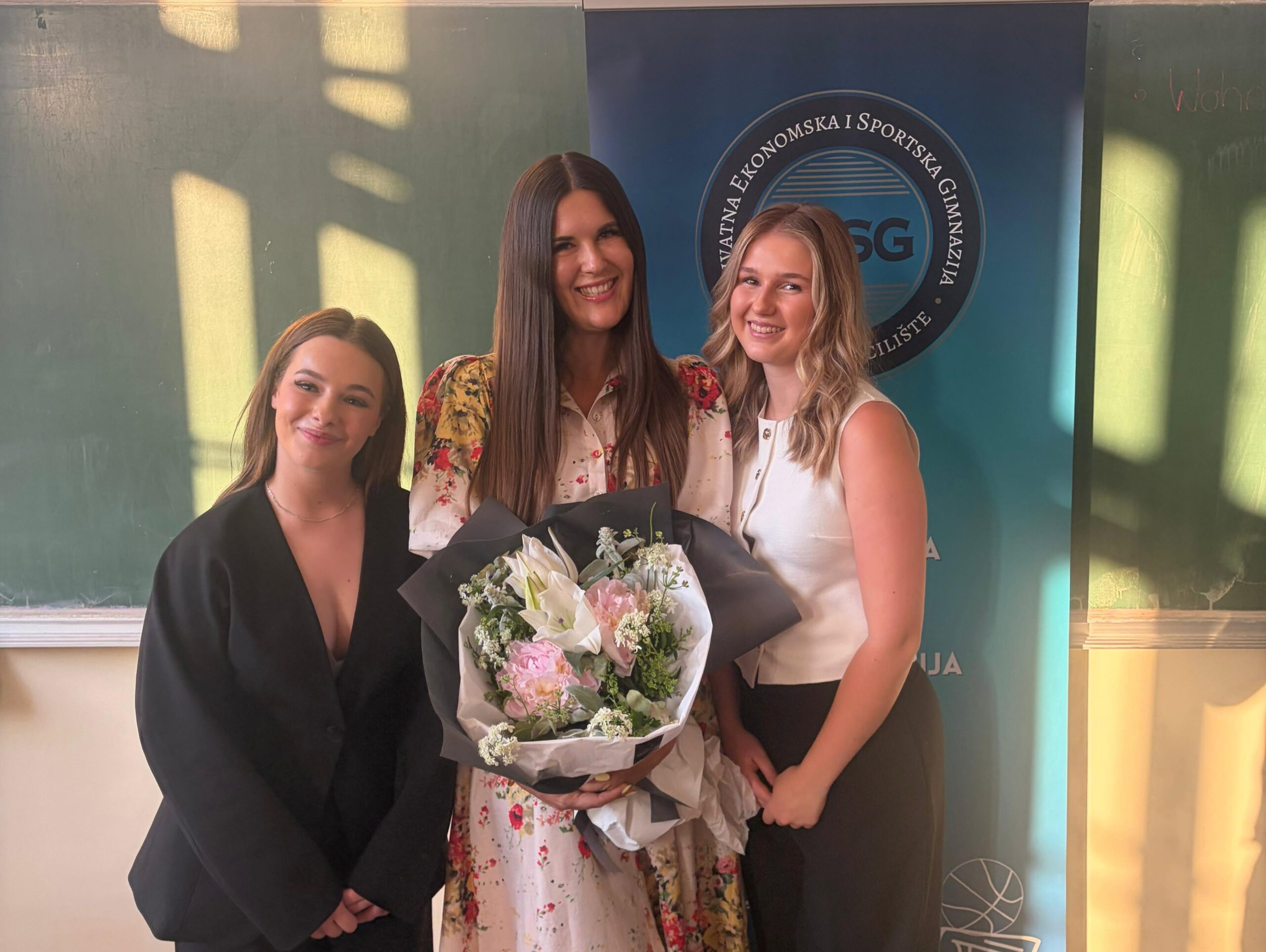 Three young women pose together at a graduation ceremony, one in a black gown holding a bouquet, with a blue banner in the background.