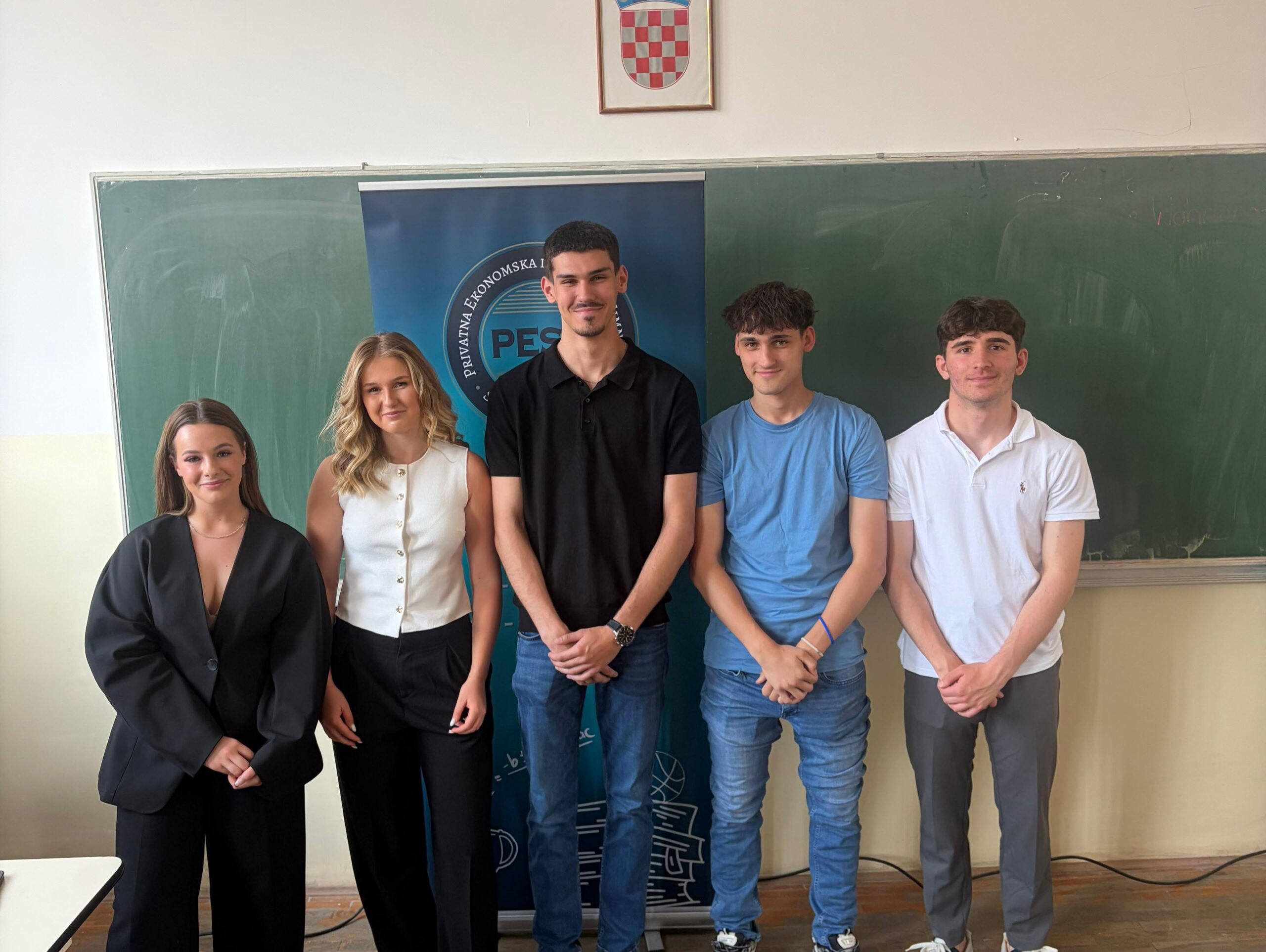 Five students posing together indoors in front of a blue banner and chalkboard, in casual/formal attire.