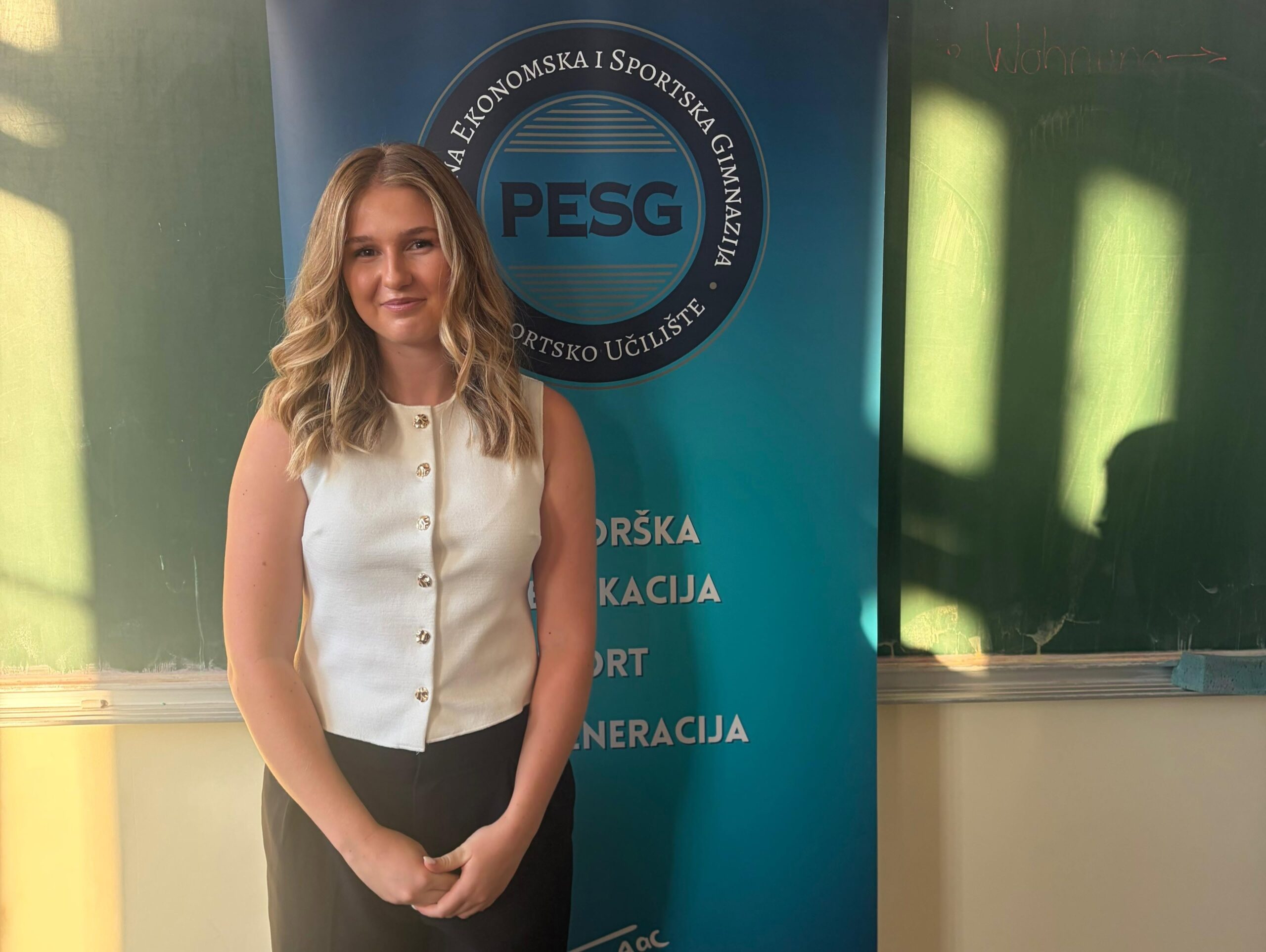 Young woman with blonde hair in a white sleeveless top stands beside a blue PESG banner at an event, smiling.