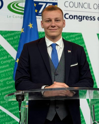 Male speaker in a dark suit with a blue tie stands behind a clear podium, hands folded, with an EU flag and conference backdrop behind him.