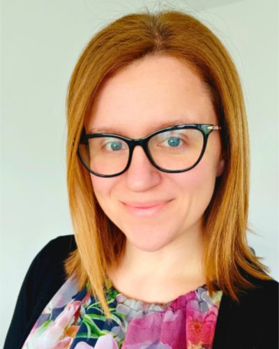 Portrait of a woman with shoulder-length red hair, black-framed glasses, and a floral blouse, smiling at the camera.