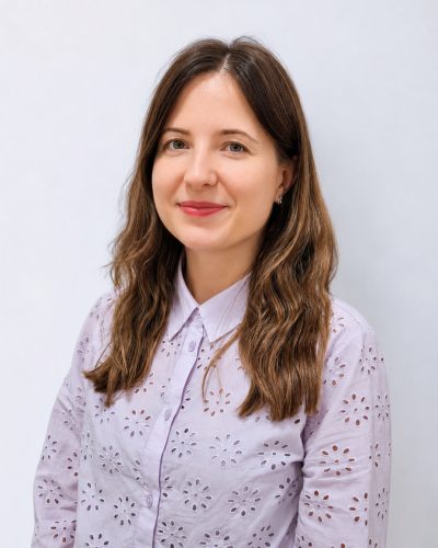 Smiling woman with long brown hair in a lavender eyelet blouse, posing against a light gray background (studio portrait).